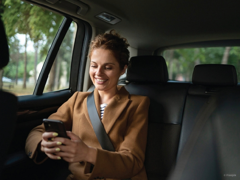 Happy woman sitting inside a car, in the backseat, holding a smartphone.