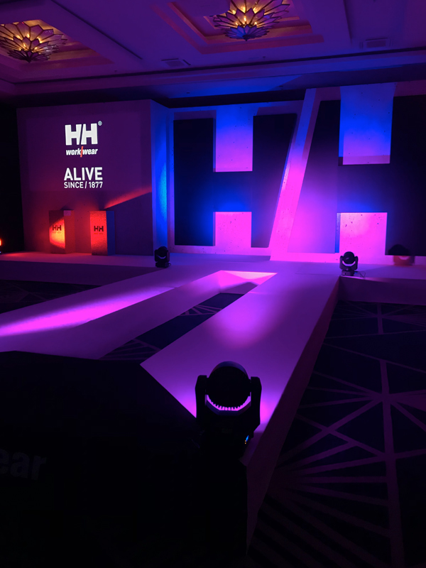 Stage illuminated with blue, purple and red lights and decorated with a logo on the wall for an indoor event.